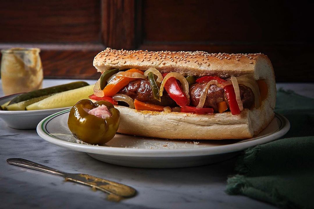 Close-up of an Italian sausage sandwich with colorful peppers, onions, and sesame seed bun on a white plate with antique silverware.