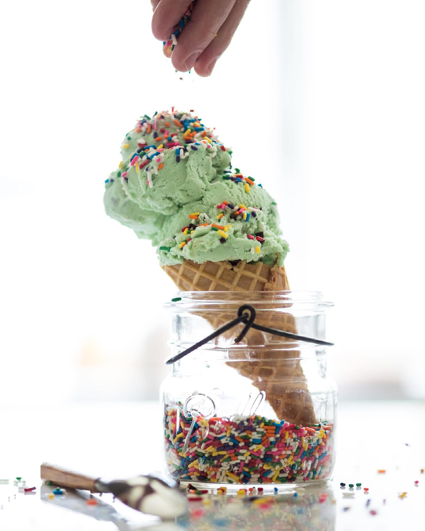 Ice cream cone in a glass jar being decorated with sprinkles by a hand in a bright, airy setting with a utensil on the table.
