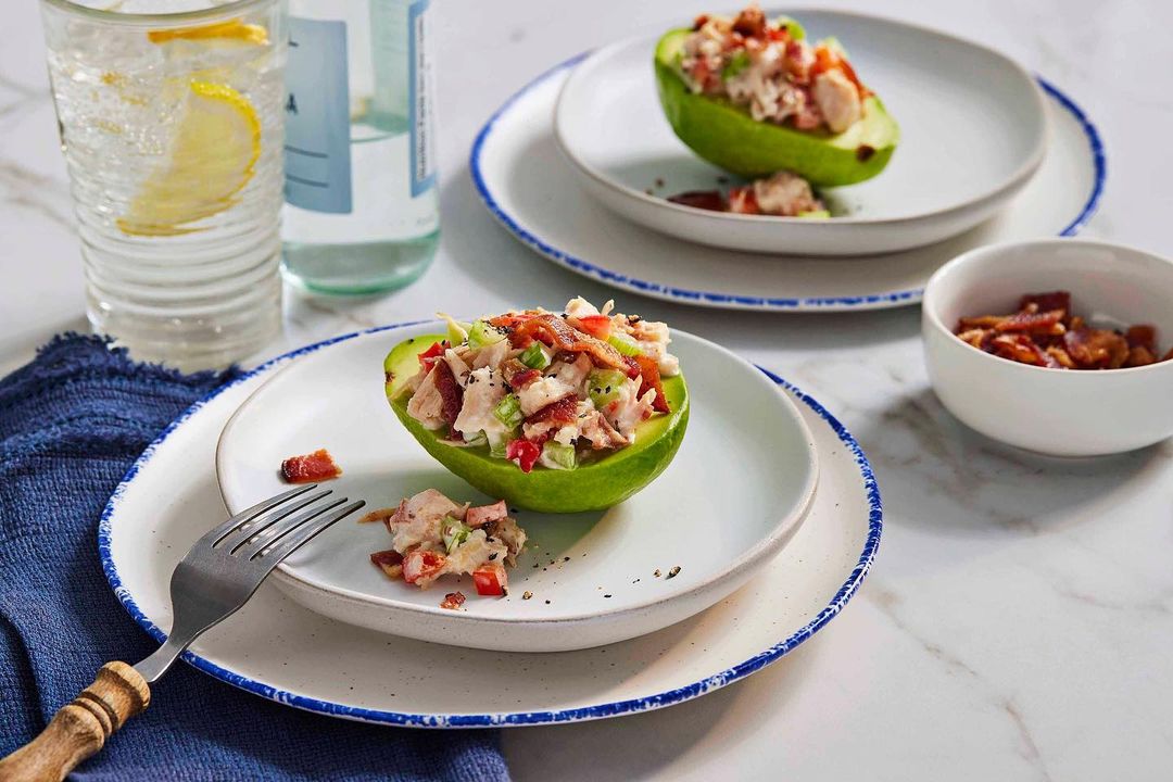 Chicken salad stuffed avocados on plates. Water glass and small bowl on white marble table with blue napkin. Food and beverage photo.