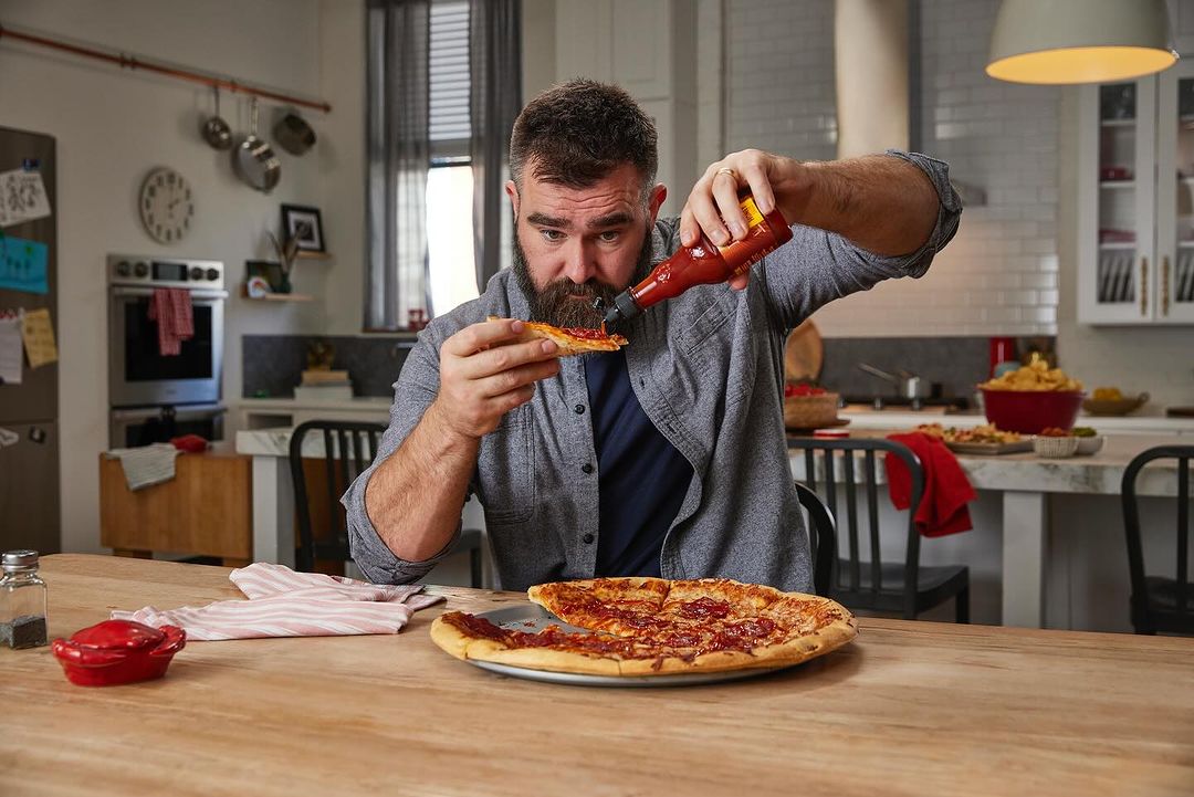 Bearded man in a kitchen putting hot sauce on a slice of pizza with a full pizza, stove, cabinets, and kitchenware in the background.