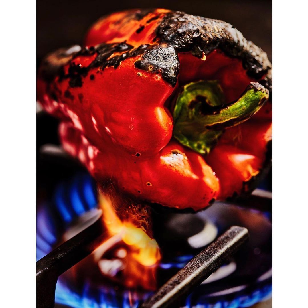 Grilled red bell pepper with charred skin and green stem, cooking on a stove burner over blue flame, food photography close-up.