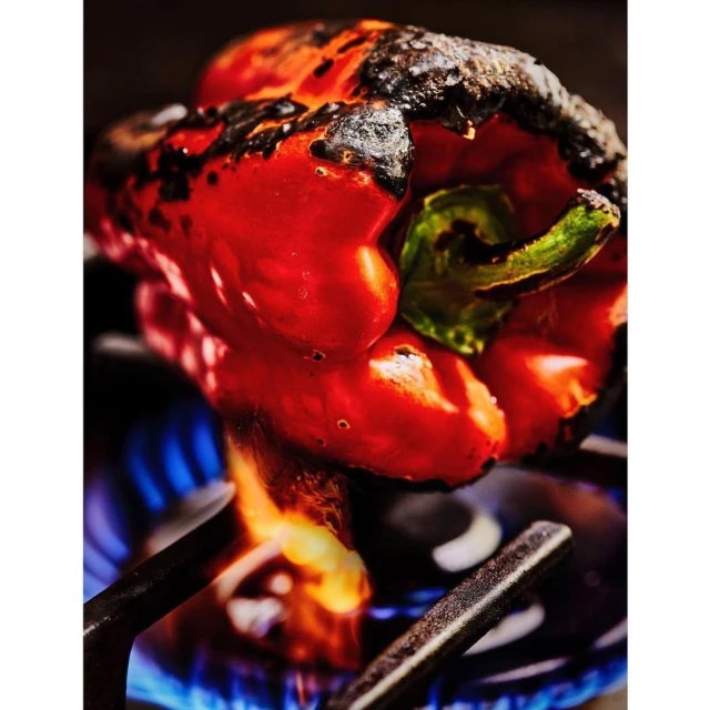 Grilled red bell pepper with charred skin and green stem, cooking on a stove burner over blue flame, food photography close-up.