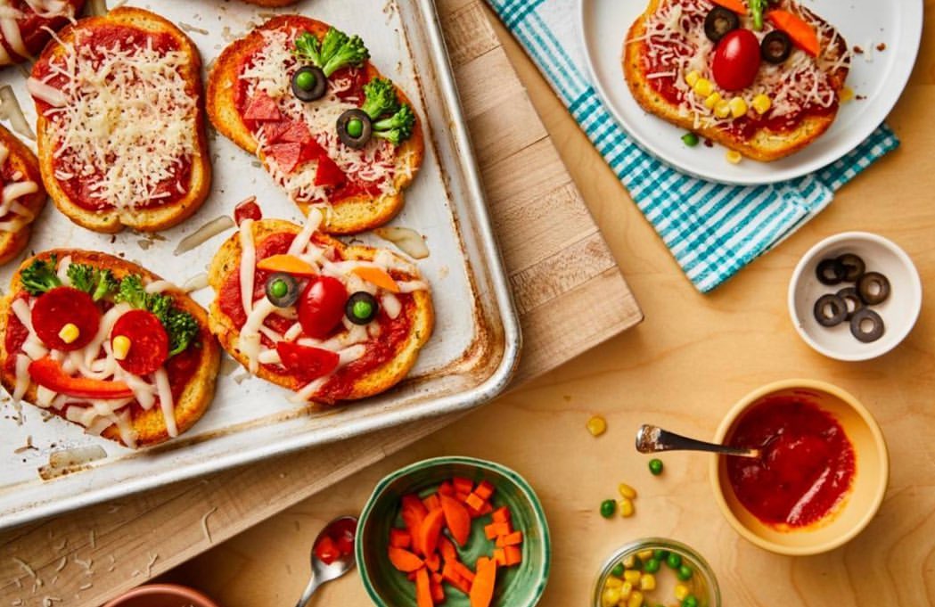 Overhead shot of toast pizzas decorated to look like faces with tomato sauce, cheese, olives, corn, broccoli, and carrots on a baking sheet.