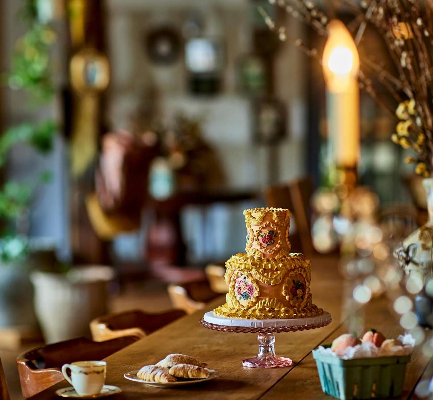 Golden tiered cake with floral accents sits on a table with croissants, peaches, and a teacup in a warm, inviting setting.