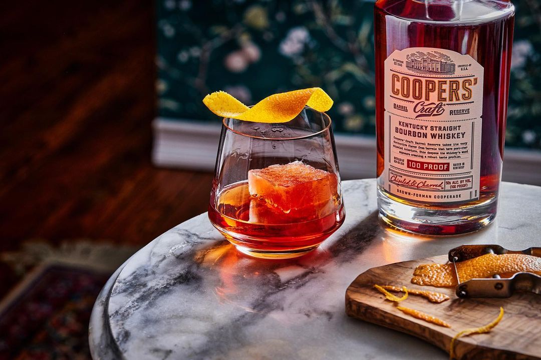 Bourbon whiskey cocktail in a glass with an ice cube and orange peel, set on a marble table next to a bottle and cutting board.
