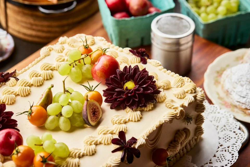Square cake decorated with fruit, including grapes and figs, sits on a wooden table next to a metal shaker and plates.