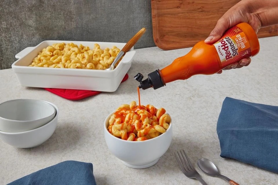 Macaroni and cheese is drizzled with buffalo sauce from a squeeze bottle, sitting on a table setting with bowls and serving utensils.