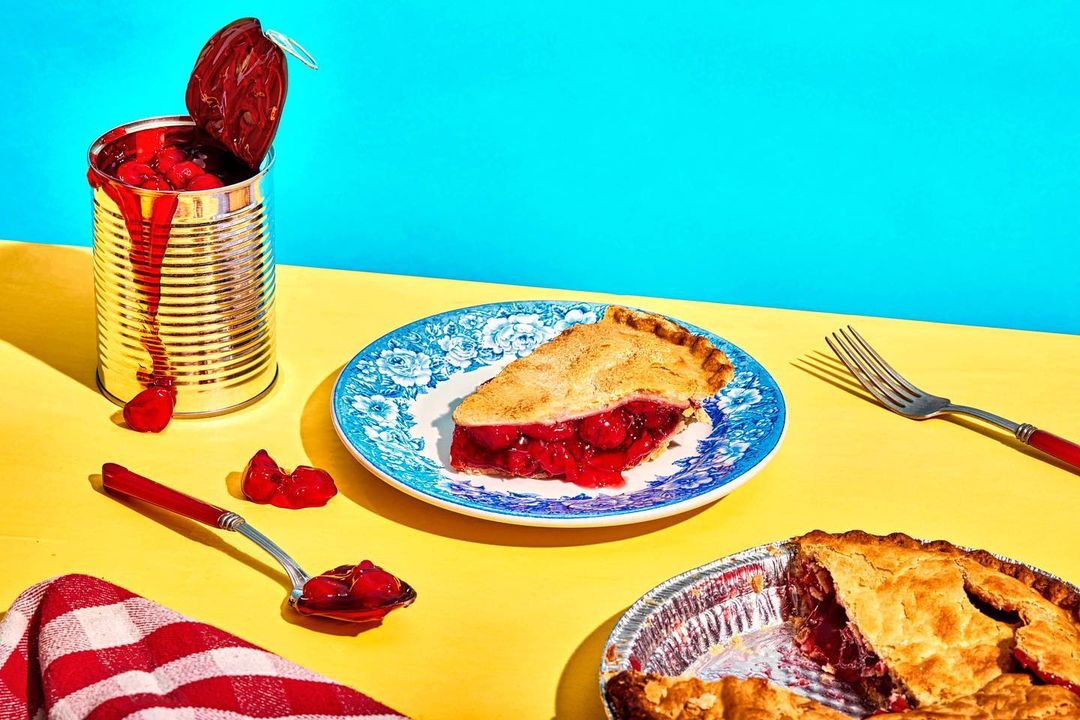Overhead shot of a slice of cherry pie on a plate, surrounded by a can of cherries, pie filling, a fork, and a red-checkered cloth.