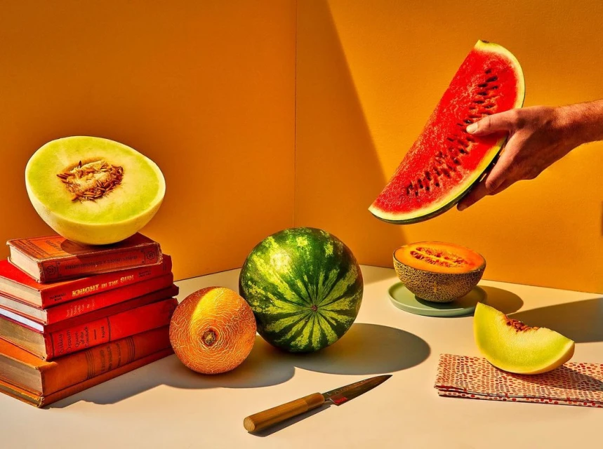 Still life photo of a hand holding a watermelon slice, various melons, a knife, and books against an orange background.