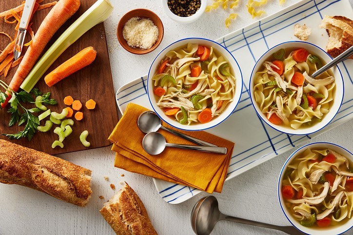 Overhead shot of three bowls of chicken noodle soup with bread, celery, and carrots on a white wooden table with a serving tray.