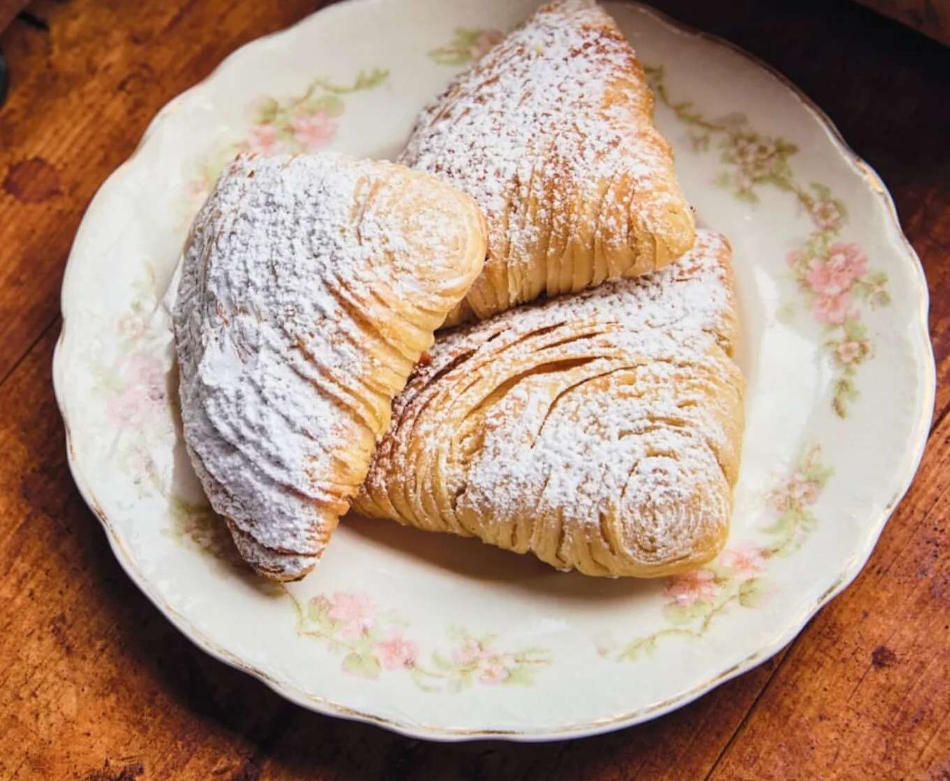Three sfogliatelle pastries dusted with powdered sugar are arranged on a decorative plate on a wooden surface, top-down view.