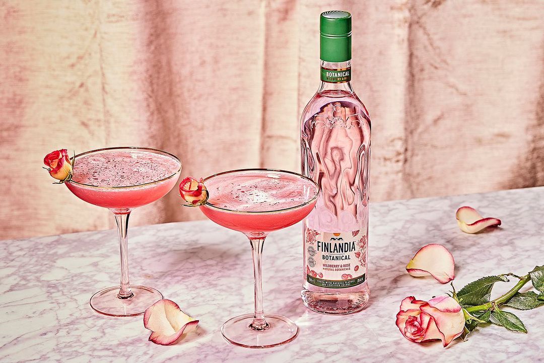 Two pink cocktails are served in coupe glasses next to a Finlandia botanical spirit bottle, with rose petals scattered across a marble table.
