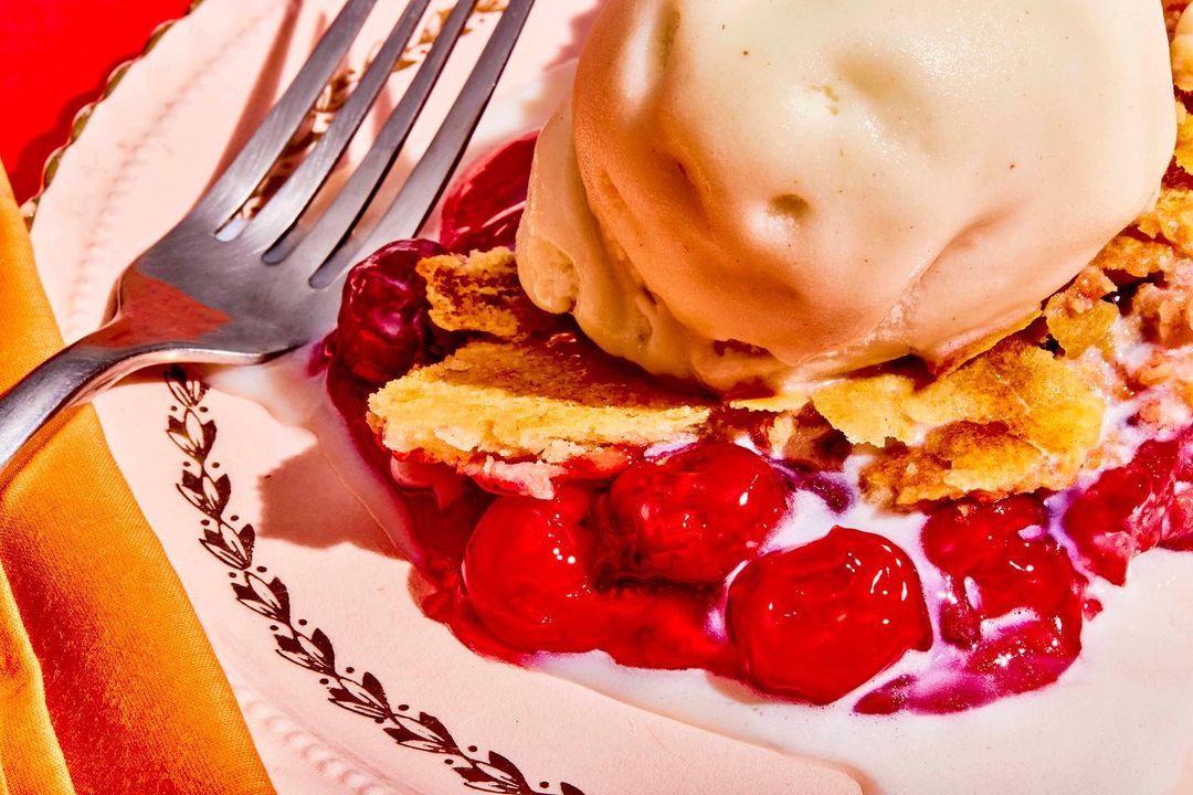 Cherry pie with vanilla ice cream sits on a decorative plate next to a metal fork and a gold ribbon, creating a dessert still life.