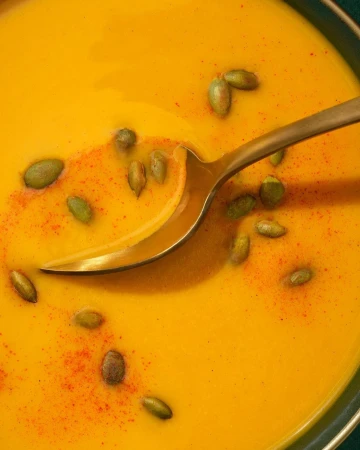 Overhead shot of golden pumpkin soup with spoon and pumpkin seeds, warm and inviting for culinary and seasonal-themed content.