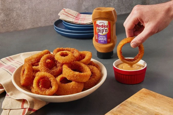 Golden-fried onion rings piled on a plate, someone dipping an onion ring into a small red bowl of creamy sauce, food photography.