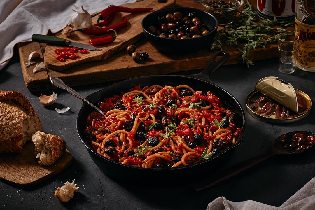 Overhead shot of spaghetti puttanesca in a cast iron pan, alongside bread, olives, anchovies, and spices on a dark surface.