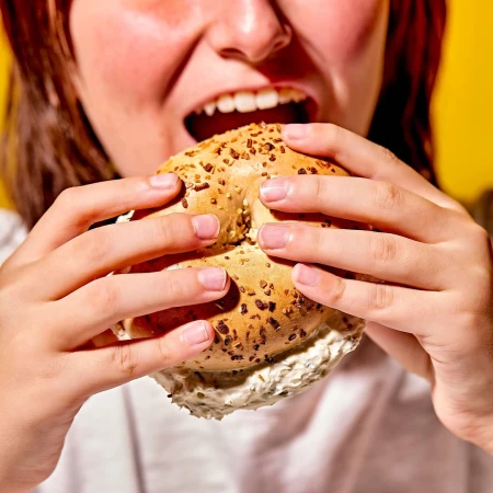 Close-up of a person holding and about to eat a bagel with cream cheese against a bright yellow background.