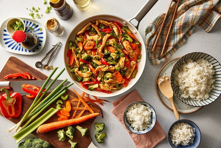 Overhead shot of chicken stir-fry with broccoli, carrots, and peppers in a white pan, surrounded by rice bowls and vegetables.