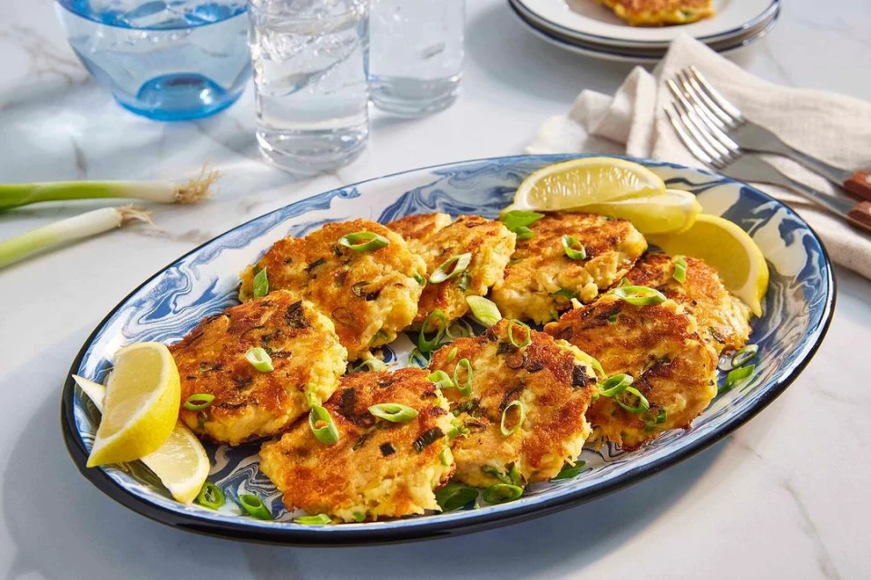 Close-up of crab cakes arranged on a blue patterned plate, garnished with scallions and lemon wedges, set on a marble surface.