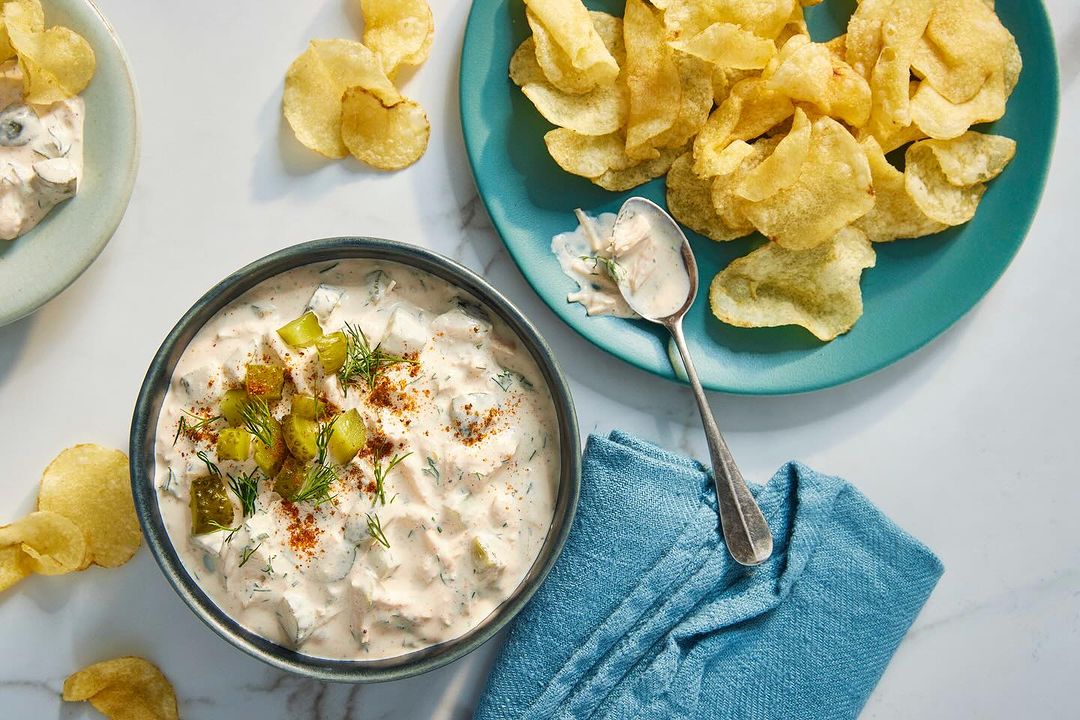 Overhead shot of dill pickle dip in a bowl with potato chips on plates, paired with a blue cloth on a white marble surface.