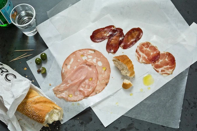 An overhead shot captures a selection of cured meats, sliced bread, olives, and a glass of water arranged on white parchment paper.