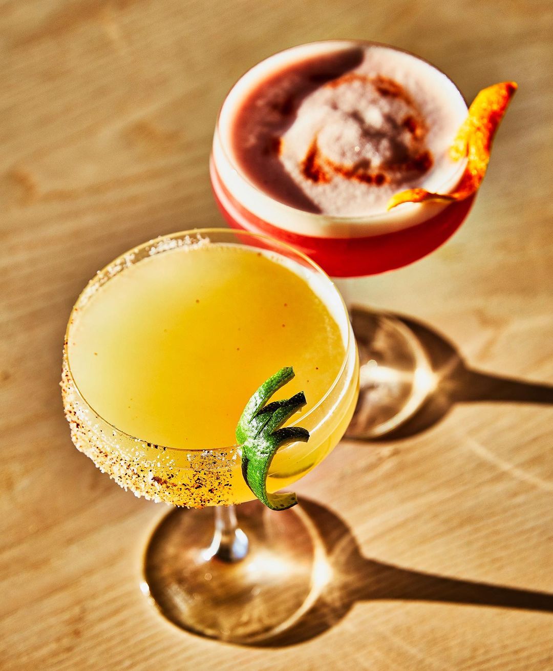 Overhead shot of two vibrant cocktails: one yellow with a green garnish and one red with a frothy top, set against a light wood surface.