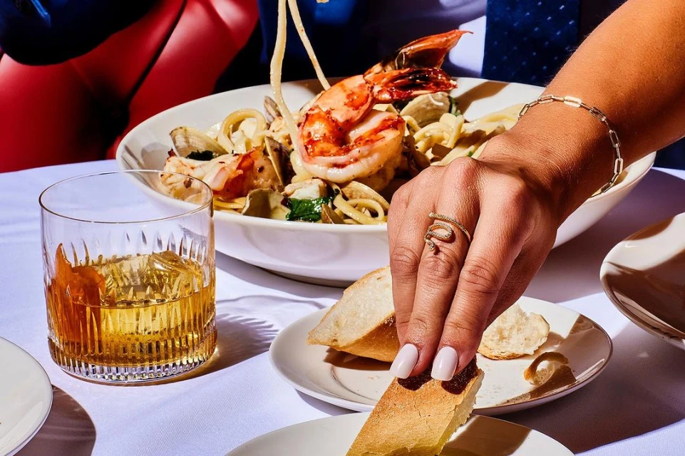 Close-up of seafood pasta with shrimp, clams, and pasta alongside a glass of whiskey, bread, and a hand with jewelry on a white tablecloth.