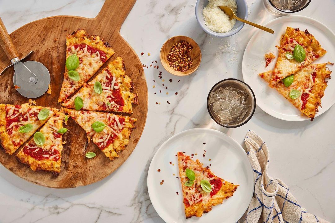 Overhead shot of a homemade pizza cut into slices, arranged on a wood cutting board with basil garnish, alongside plates and glasses.