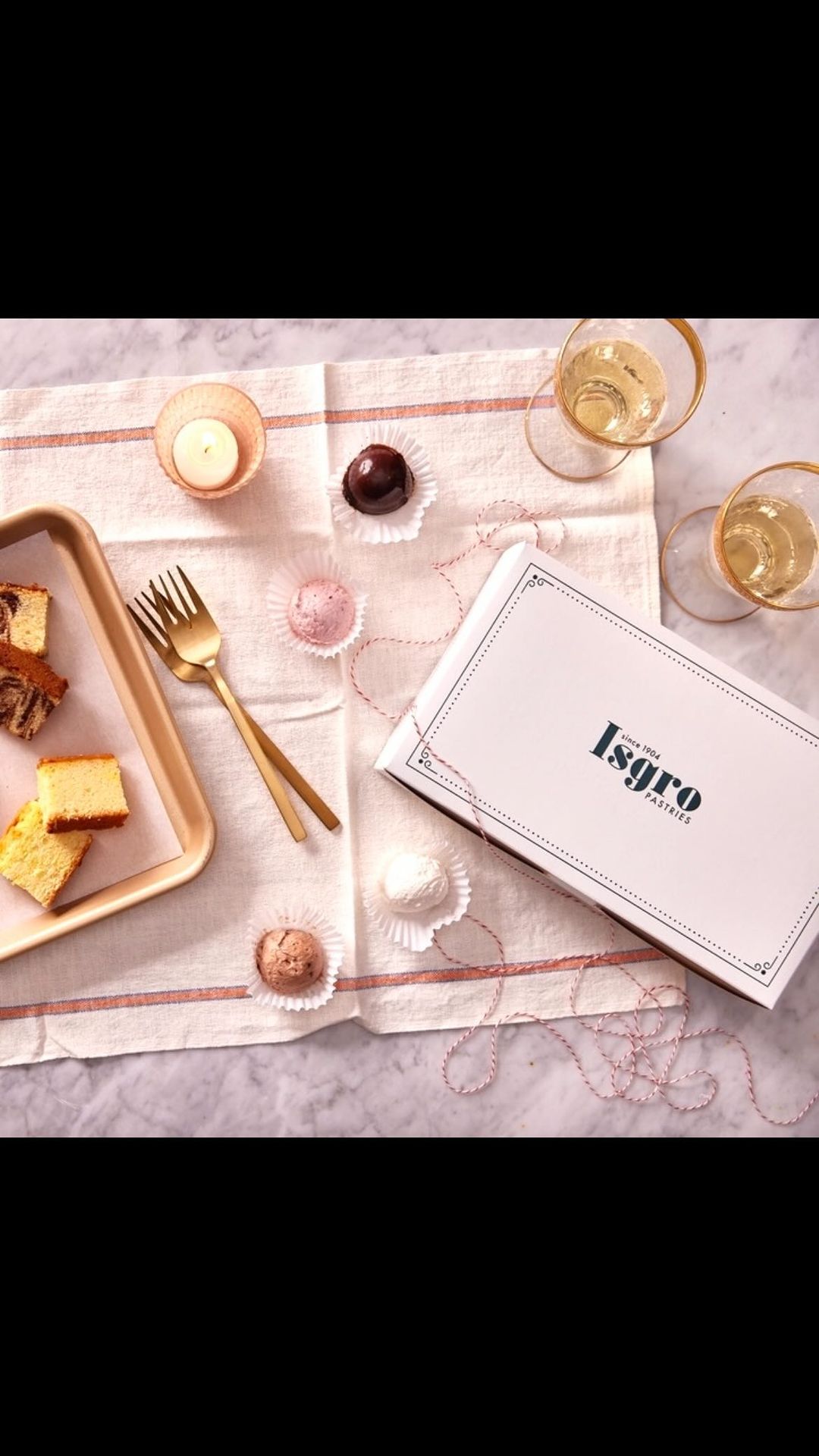 Overhead shot of cake slices, chocolate truffles, champagne glasses, and a bakery box on a striped linen tablecloth.