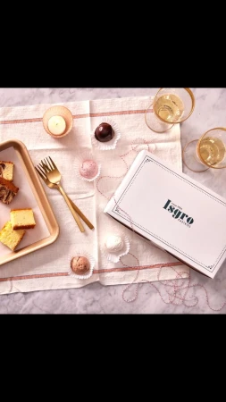 Overhead shot of cake slices, chocolate truffles, champagne glasses, and a bakery box on a striped linen tablecloth.