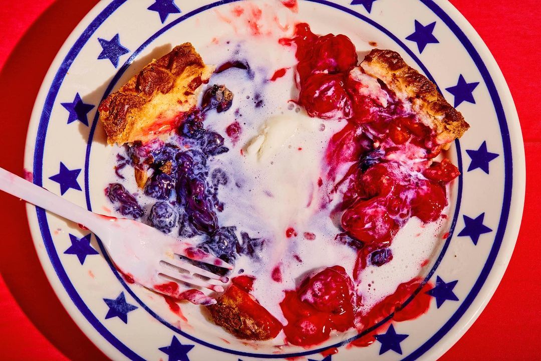 Overhead shot of a plate with remnants of berry pie, ice cream, and a plastic fork, set against a bright red background.