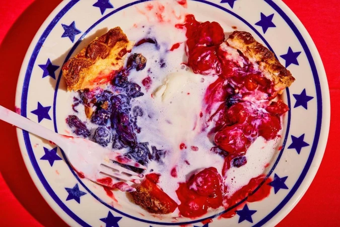Overhead shot of a plate with remnants of berry pie, ice cream, and a plastic fork, set against a bright red background.