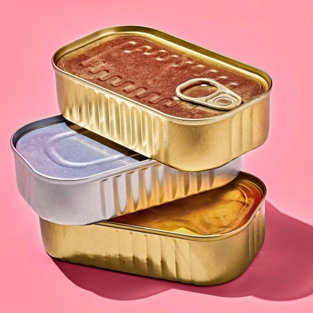 A stack of three rectangular metal tin cans, showcasing silver and gold colors, set against a plain pink background.