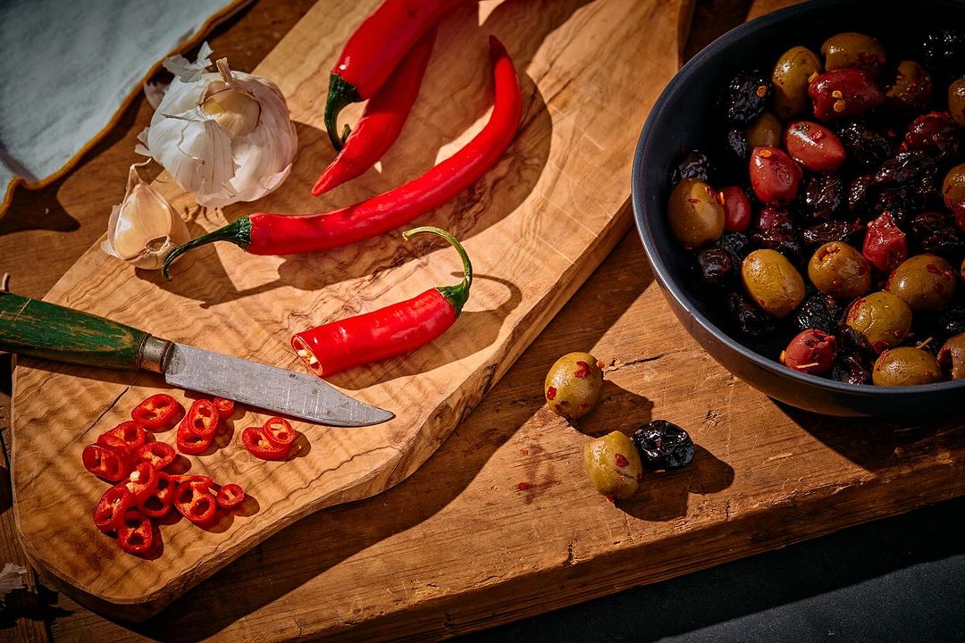Still life composition with red chili peppers, olives in a bowl, garlic bulb, and a knife on a wooden cutting board.