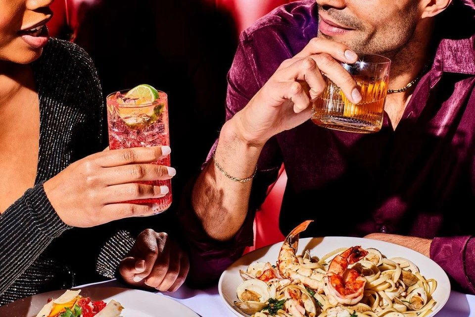 A couple seated at a table with plates of seafood pasta and cocktails, the dark-haired man holds a whisky glass with amber liquid.
