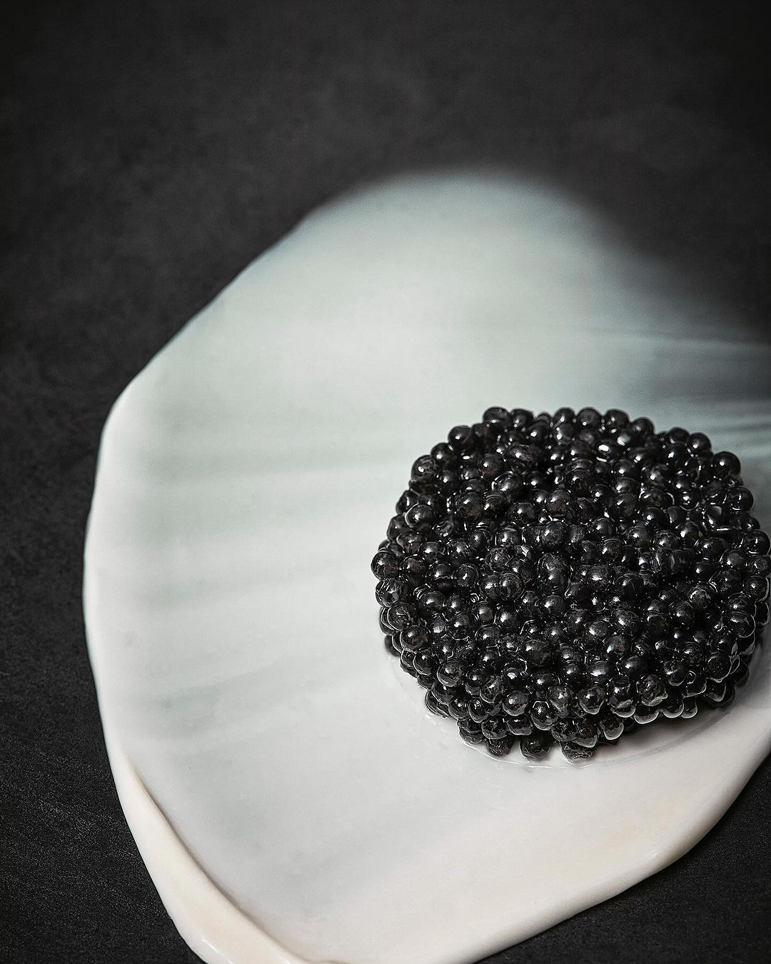 Close-up shot of black caviar arranged on an off-white curved plate against a dark background, food photography, studio lighting.