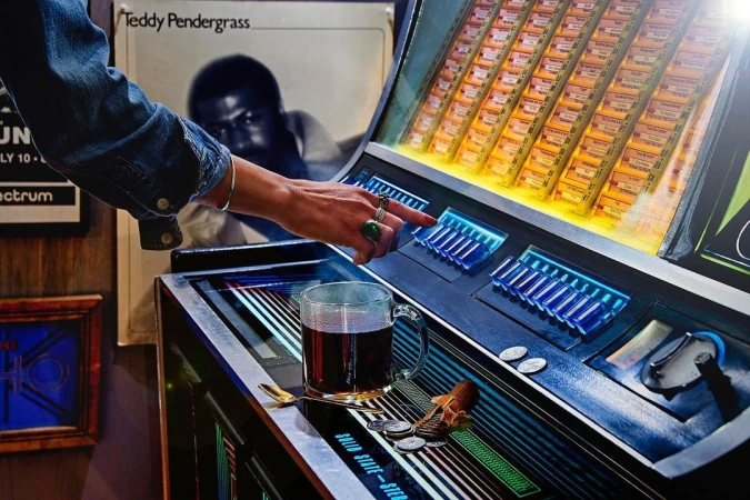 Woman selects music on vintage jukebox with vinyl record cover, drinking glass, spoon, and coins on surface in dimly lit interior.