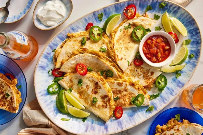 Overhead shot of quesadillas on a blue-patterned platter with limes, salsa, hot peppers, sour cream, and orange soda. Food photography.