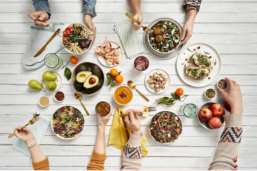 Overhead shot of a white table covered with an array of healthy dishes, fruits, and beverages, surrounded by people's hands and arms.