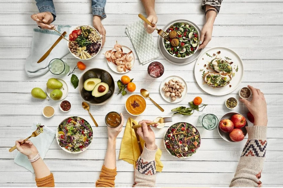 Overhead shot of a white table covered with an array of healthy dishes, fruits, and beverages, surrounded by people's hands and arms.