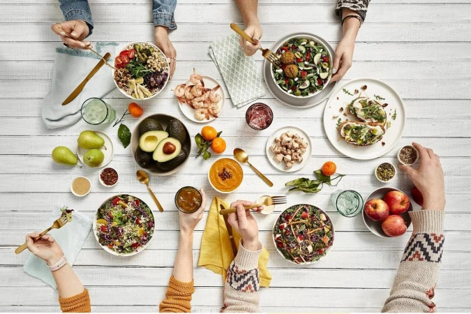 Overhead shot of a white table covered with an array of healthy dishes, fruits, and beverages, surrounded by people's hands and arms.