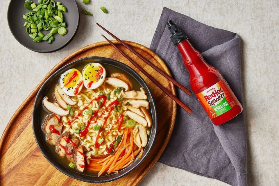 Overhead shot of ramen bowl on wooden tray with hard-boiled egg, sliced mushrooms and carrots, with a red squeeze bottle.