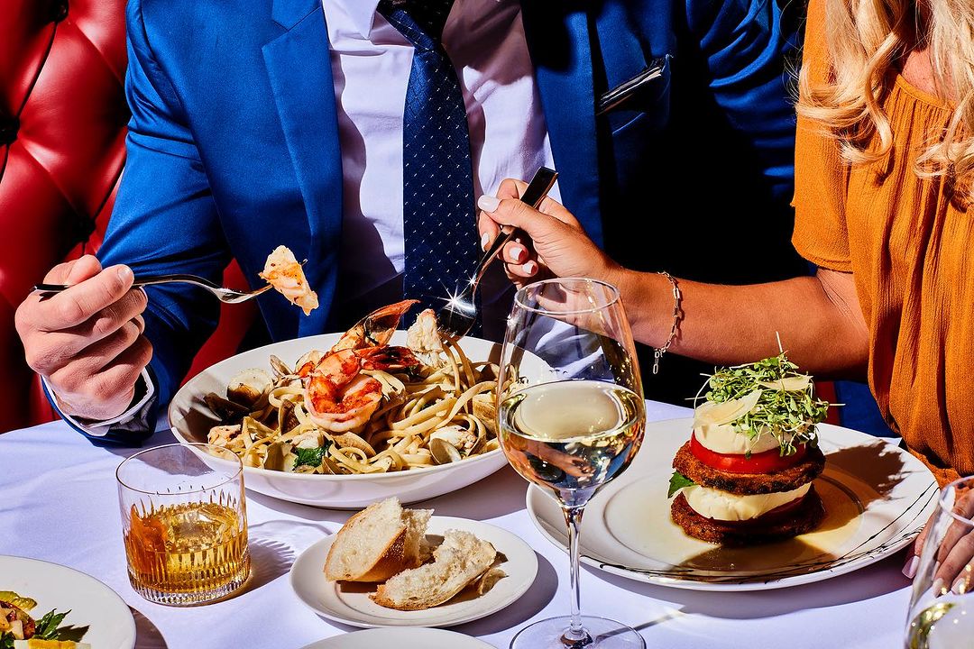 Overhead shot of two people dining at a restaurant, enjoying pasta with seafood, a glass of wine, and a stacked appetizer.