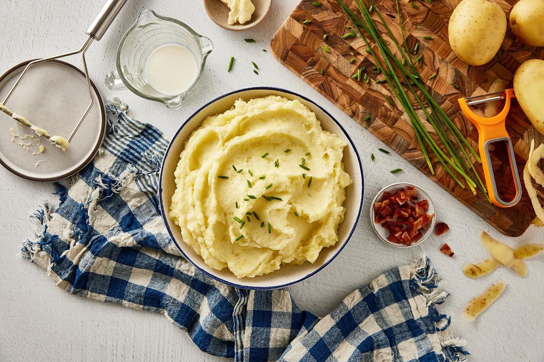 Overhead shot of mashed potatoes in a bowl, garnished with chives, surrounded by ingredients and utensils on a white surface.