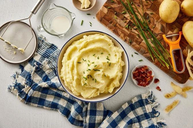 Overhead shot of mashed potatoes in a bowl, garnished with chives, surrounded by ingredients and utensils on a white surface.