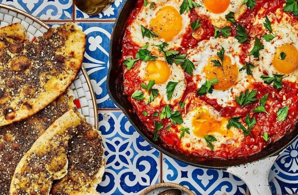 Overhead shot of shakshuka in a cast iron pan with za'atar bread, featuring a vibrant blue and white patterned tile backdrop.