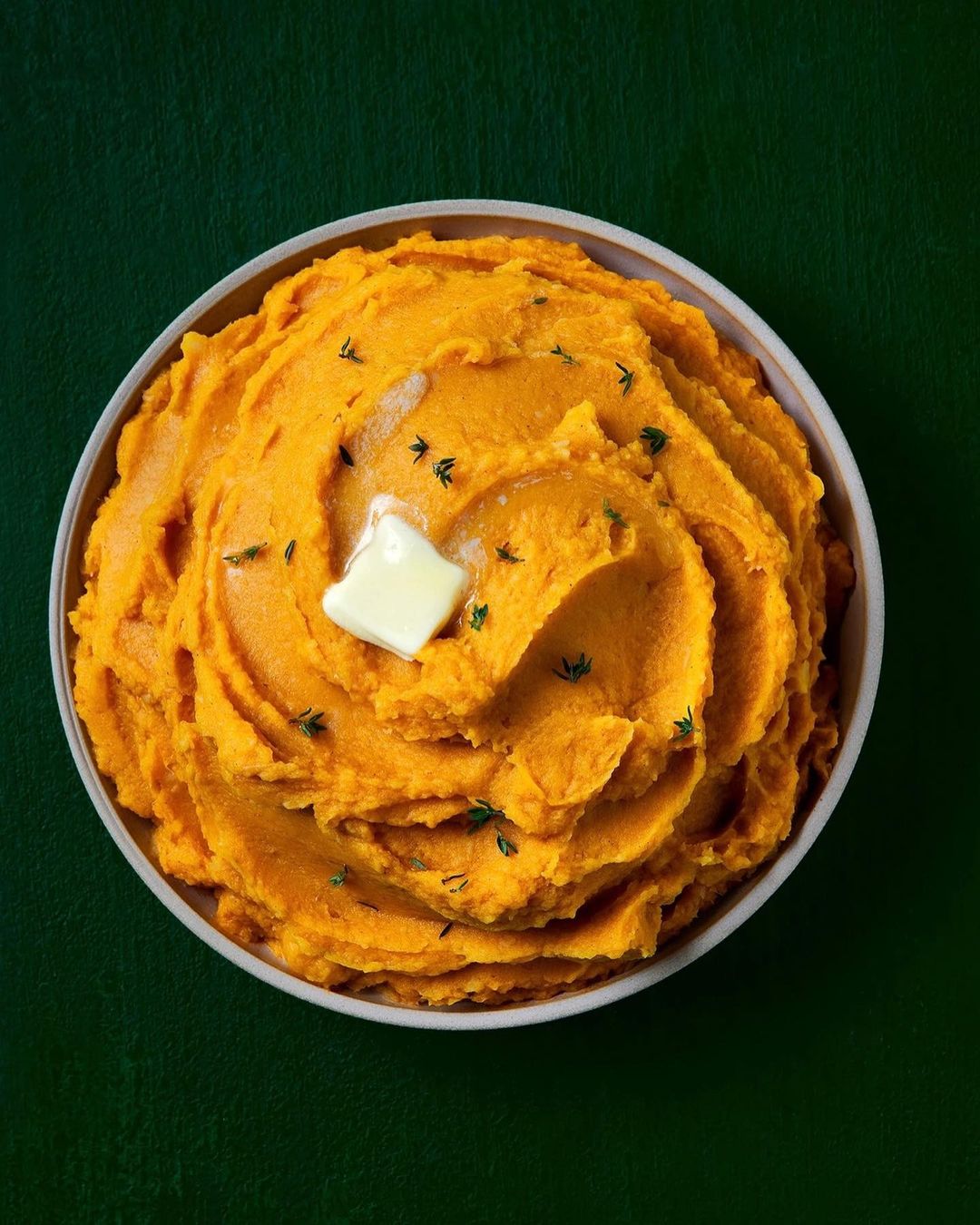 Overhead shot of mashed sweet potatoes in a white bowl, topped with butter and herbs on a dark green background.