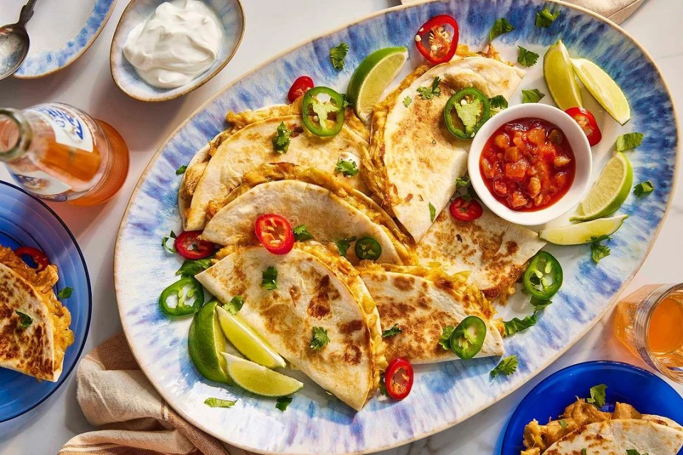 Overhead shot of quesadillas arranged on a platter with salsa, lime, peppers, hot sauce, and sour cream creating a delicious food spread.