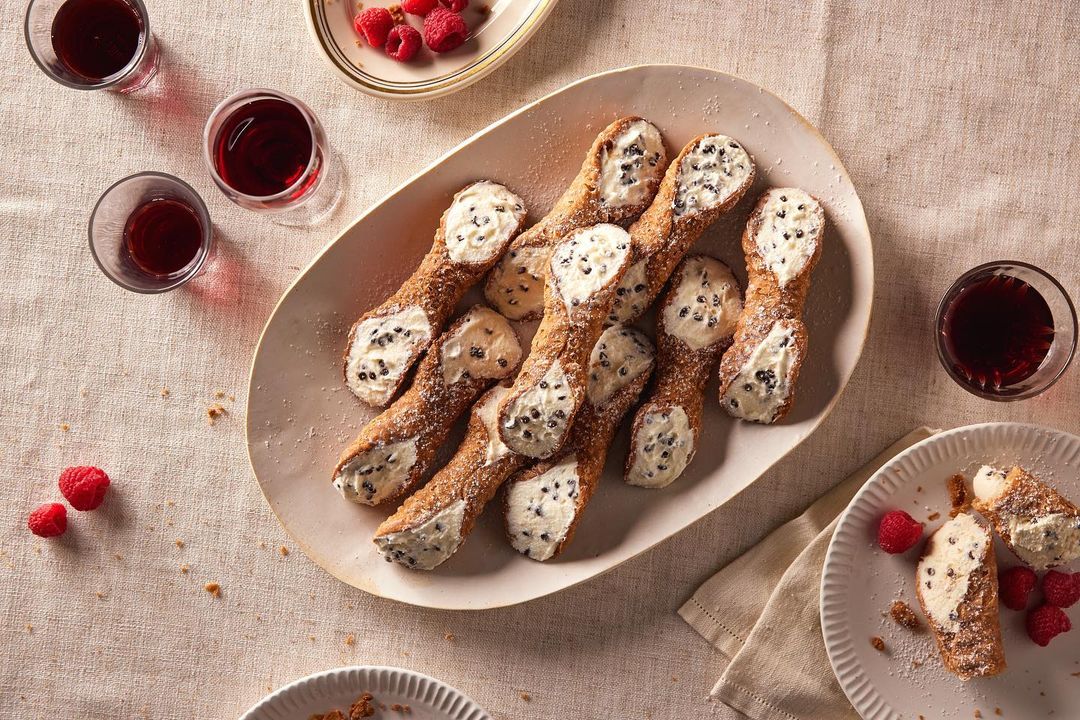 Overhead shot of cannoli pastries filled with cream and chocolate chips on a platter, alongside glasses of red wine and fresh raspberries.