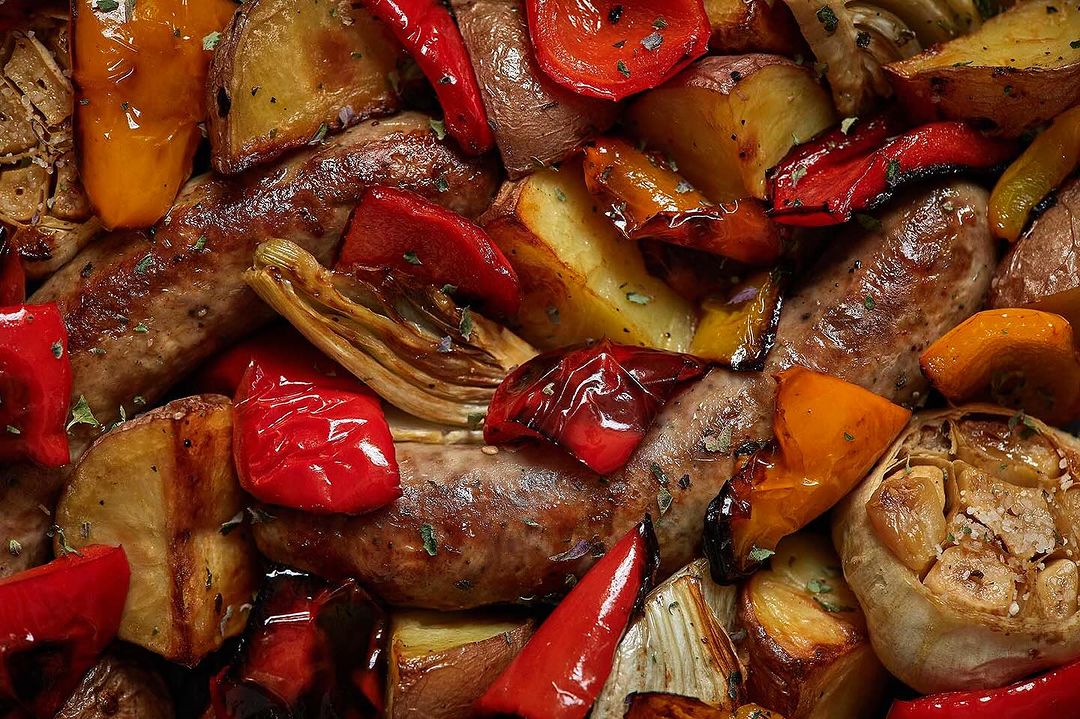 Overhead shot of a dish with sausage, roasted potatoes, red bell peppers, yellow bell peppers, and roasted garlic.